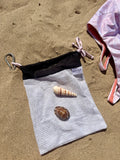 A white mesh beach bag with a black top band and metal carabiner, laid on sandy ground with two seashells placed inside. A pink swimsuit edge is visible at the right edge.